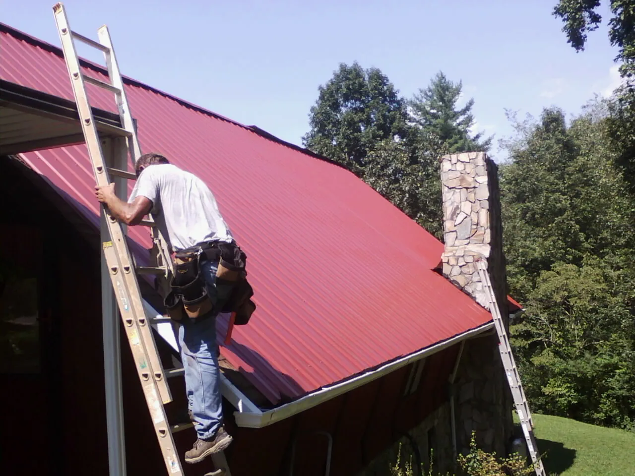 Roofer on a ladder beside a red metal roof and stone chimney — Soffit & Fascia Repair by Roofing Brothers in Brandeis